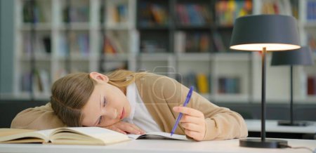 Photo for Sad school girl lying on book and entertaining herself by playing with pen, pupil at boring lesson feeling tired or depressed, bookshelves on blurred background. Concept of education - Royalty Free Image