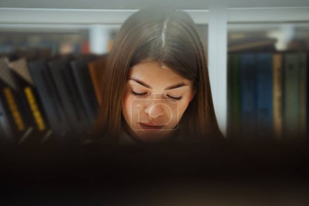 Photo for Young woman with freckled face and long hair reading book, peeped from behind bookshelves. Handheld reader enjoying interesting literature in library. Concept of education - Royalty Free Image