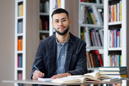 Photo for Young bearded male student with eyeglasses wearing jacket sitting in library and making notes on paper, looking up at camera and smiling. Portrait of intelligent man studying. Concept of education - Royalty Free Image