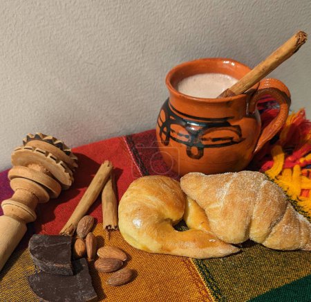 Mexican bread, chocolate pieces, almonds and cinnamon stick. Wooden mortar and clay mug with hot chocolate on colorful mexican tablecloth