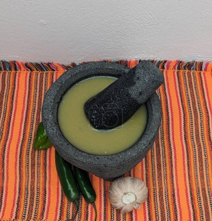 Foto de Mortero de piedra mexicano y mortero con salsa verde, ajo, chiles sobre mantel mexicano de naranja - Imagen libre de derechos
