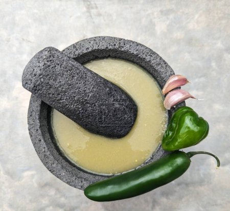 Foto de Mortero y mortero de piedra, salsa verde mexicana, dientes de ajo, habanero y chiles jalapeños. Posición lateral sobre fondo de cemento - Imagen libre de derechos