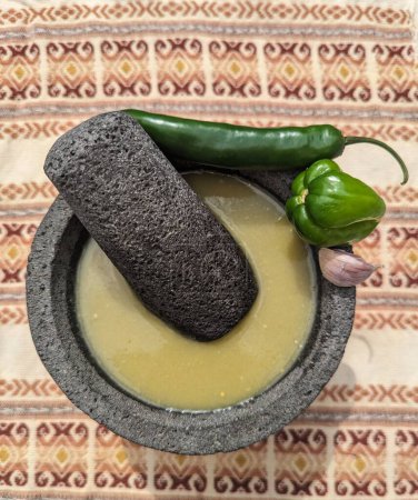 Foto de Mortero de piedra mexicano y mortero con salsa verde, ajo, chiles sobre mantel mexicano de naranja - Imagen libre de derechos