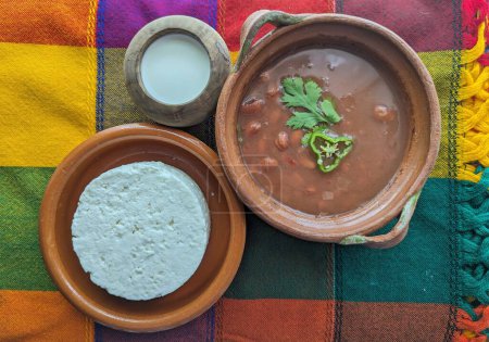 Mexican clay cooking pot with beans, cilantro and chili. Cream and panela cheese on traditional tablecloth