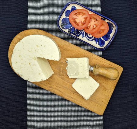 Sliced panela cheese on wooden dish. Sliced tomato in ceramic recipient. Blue tablecloth