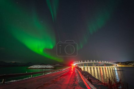 Foto de Hermosas luces boreales sobre el puente iluminado de Sommary con el semáforo rojo. bailando Aurora boreal reflejándose en el agua del Atlántico Norte. luz verde sobre la isla frente a Troms - Imagen libre de derechos