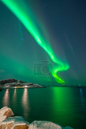 Foto de Hermosas luces del norte sobre una casa iluminada en el pueblo Brensholmen en la isla de Kvaloya cerca de Tromso. bailando luces polares sobre una colina, luz verde en el cielo en invierno, en el agua reflejando luces de Aurora Boreal en Noruega - Imagen libre de derechos