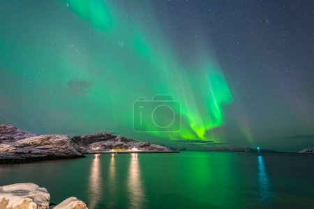 Foto de Hermosas luces del norte sobre una casa iluminada en el pueblo Brensholmen en la isla de Kvaloya cerca de Tromso. bailando luces polares sobre una colina, luz verde en el cielo en invierno, en el agua reflejando luces de Aurora Boreal en Noruega - Imagen libre de derechos