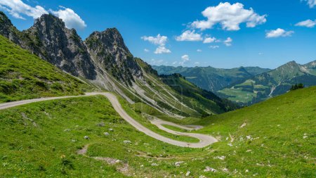 Téléchargez les photos : Une étroite route de montagne serpente vers le bas de la montagne dans le Grosswalsertal, le long des montagnes pierreuses sur les prairies fleuries escarpées dans le Vorarlberg, Autriche - en image libre de droit