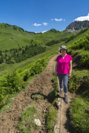 Foto de Mujer senderismo en el camino a través de la hermosa pradera de montaña llena de flores de colores. El excursionista pasa por el gran valle de Walser con la montaña llamada pared roja en el fondo, hermoso paisaje alpino - Imagen libre de derechos