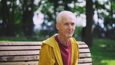 Téléchargez les photos : Homme âgé souriant assis sur le banc après l'entraînement dans le parc, se reposant après l'entraînement - en image libre de droit