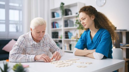 Rehabilitation doctor watching old woman connecting jigsaw pieces, brain teaser
