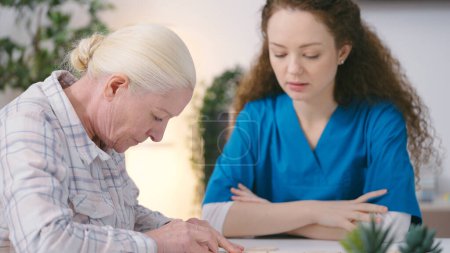 Freundliche Krankenschwester beobachtet Seniorin beim Verbinden von Puzzleteilen, Therapiesitzung