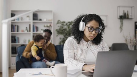 Téléchargez les photos : Père joue avec son enfant pendant que maman porte des écouteurs et travaille sur son ordinateur portable - en image libre de droit