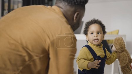 Téléchargez les photos : Bébé afro-américain marchant de façon instable, souriant père regardant les pas de bébé - en image libre de droit