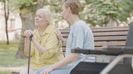 Téléchargez les photos : Bénévole utile soutenant la vieille femme triste assise sur le banc avec un bâton de marche - en image libre de droit