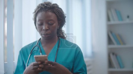 Téléchargez les photos : Infirmière afro-américaine sérieuse composer le numéro sur son téléphone portable, appeler les parents du patient pour informer sur le bien-être - en image libre de droit