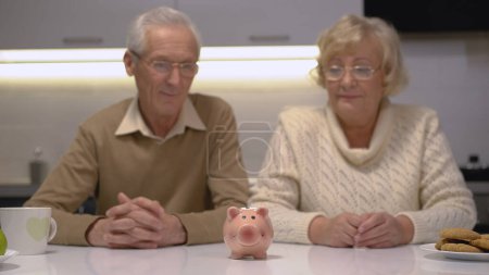 Photo for A senior couple looking at a piggy bank standing on the table while planning their family budget - Royalty Free Image