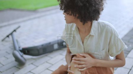 Traurige lockige Frau berührt schmerzende Knie, sitzt auf dem Boden in der Nähe von Elektroroller, Unfall