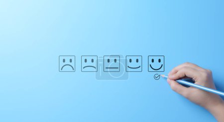Photo for Customer Satisfaction Rating Scale with Smiley Faces. A hand marks a happy face on a customer satisfaction survey with various emoticon options ranging from sad to happy on a blue background. - Royalty Free Image