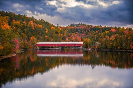 The Wakefield Gendron Bridge over the Gatineau River during autumn, colorful fall foliage, dramatic stormy sky, landscape and sky reflecting on water, La Peche, Quebec, Canada October 2022.