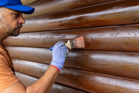Photo for Worker applying stain with a brush on wooden house exterior wall. - Royalty Free Image