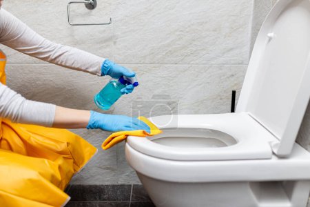 Großaufnahme weiblicher Hände in Gummihandschuhen, die Toilettenschüssel mit Desinfektionsmittel und Tuch reinigen.
