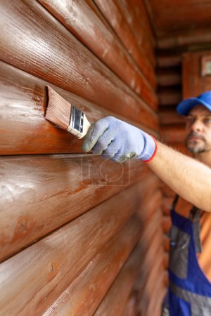 Wooden house maintenance concept. Man staining wall siding trunk.
