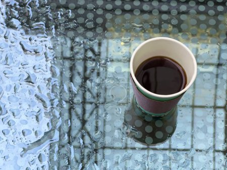 Foto de La taza de café desechable se encuentra sobre una mojada de la mesa de cristal de lluvia. En el reflejo se puede ver el cielo y la azotea de la terraza. Gotas de agua crean un patrón interesante - Imagen libre de derechos