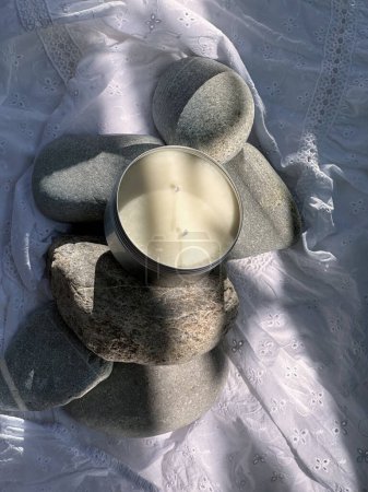 Foto de Vela en un frasco de metal redondo en color plata. Piedras y tela arrugada blanca como fondo con rayo de sol. Bodegón - Imagen libre de derechos