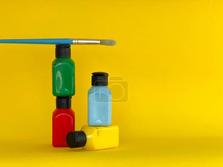 Foto de Cuatro frascos con pintura acrílica multicolor brillante y pincel sobre un fondo amarillo. Verde, azul, rojo, amarillo - Imagen libre de derechos