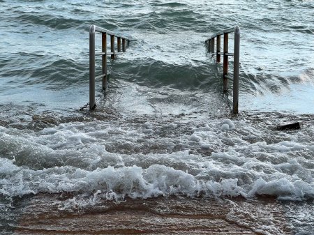 Foto de Pasarela de madera que va hacia el mar inundado. Marea, tormenta, nivel del agua elevado. Día nublado, olas activas - Imagen libre de derechos