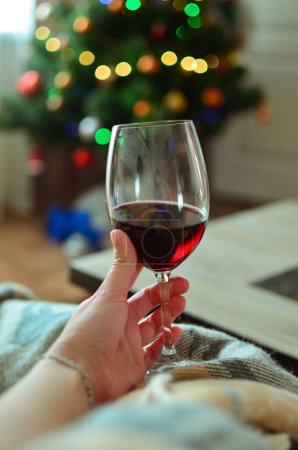 Photo for Woman holding red wine on a sofa.A glass of wine on the background of garlands and bokeh effect. A glass of red wine.Tasting. Home comfort.Christmas atmosphere with garlands and Christmas tree.Woman with a glass of wine.holds a glass in his hand. - Royalty Free Image