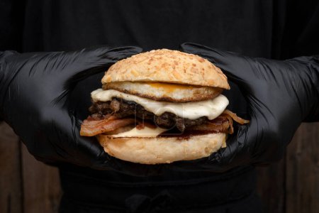 Téléchargez les photos : Chef portant des gants noirs tenant un hamburger fait de pain, œuf grillé, viande, fromage provolone et bacon croustillant. - en image libre de droit