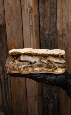 Foto de Chef con guantes negros presentando un sándwich de carne hecho con pan de ciabatta, carne, queso mozzarella, chucrut y mostaza con fondo de madera. - Imagen libre de derechos