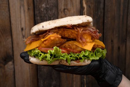 Foto de Chef con guantes sosteniendo un sándwich gourmet hecho con pan brioche, pechuga de pollo frito, lechuga, tomate, tocino y queso cheddar. - Imagen libre de derechos