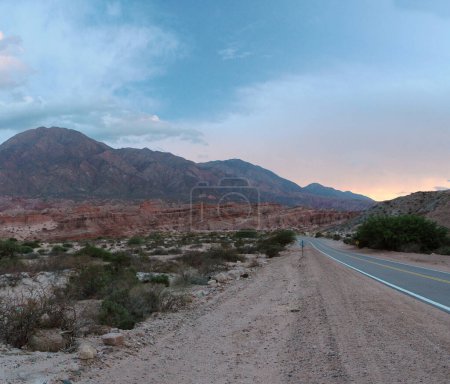 The desert at sunset. View of the desert flora, colorful mountains, sand, rock formations and empty asphalt highway across the arid valley under a beautiful sky with dusk colors.