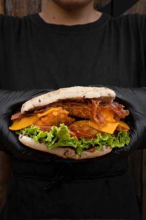 Téléchargez les photos : Chef portant des gants tenant un sandwich fait de pain brioché, poitrine de poulet frit, laitue, tomate, bacon et fromage cheddar. - en image libre de droit