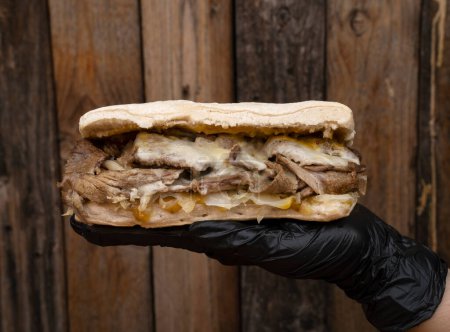 Photo for Chef wearing black gloves presenting a meat sandwich made with ciabatta bread, meat, mozzarella cheese, chucrut and mustard with a wooden background. - Royalty Free Image