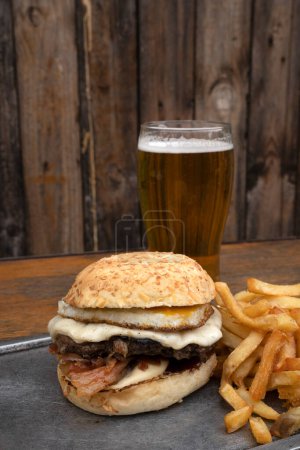 Téléchargez les photos : Burger et bière. Vue rapprochée d'un hamburger avec pain, œuf grillé, viande, fromage provolone, bacon croustillant et une pinte de bière. - en image libre de droit