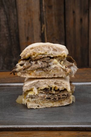Foto de Vista de cerca de un sándwich en rodajas hecho con pan de ciabatta, carne, queso mozzarella, chucrut y mostaza en un plato de metal con un fondo de madera. - Imagen libre de derechos