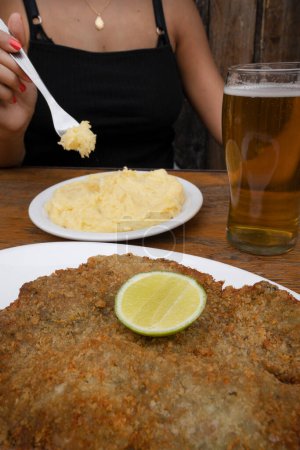 Téléchargez les photos : Vue rapprochée d'une femme qui mange une milanesa et de la purée de pommes de terre avec un verre de bière dans le restaurant. - en image libre de droit