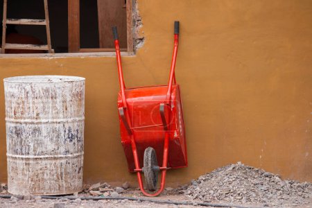 Téléchargez les photos : Site de construction. Vue d'une brouette rouge adossée au mur de béton, d'une boîte métallique et de quelques pierres, sur le chantier. - en image libre de droit