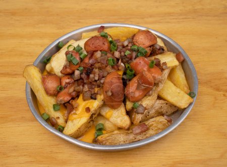 Photo for Close-up view of fried potatoes with sliced green onion, smoked sausages, crispy bacon and cheddar cheese sauce, in a metal dish on the wooden table. - Royalty Free Image