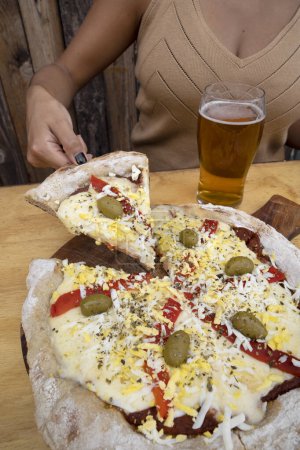 Foto de Vista de cerca de una mujer tomando una pinta de cerveza y sirviendo una rebanada de pizza con queso mozzarella, pimiento rojo asado, huevo, jamón y aceitunas verdes. - Imagen libre de derechos
