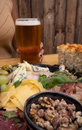 Foto de Vista de cerca de una picada tradicional, con jamón, queso, salame, aceitunas, nueces y pan focaccia. Una mujer sosteniendo un vaso de cerveza en el fondo. - Imagen libre de derechos