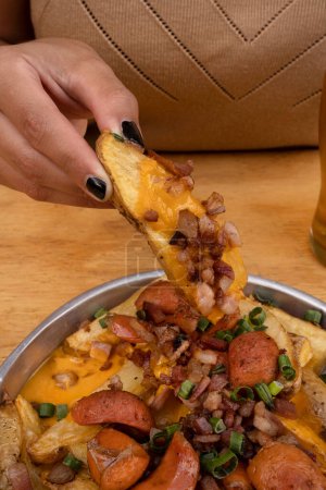 Photo for Close-up view of fried potatoes with sliced green onion, smoked sausages, crispy bacon and cheddar cheese sauce, in a metal dish on the wooden table. - Royalty Free Image