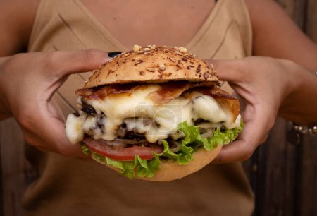 Téléchargez les photos : Burger géant. Vue rapprochée d'un délicieux hamburger au fromage, laitue, tomate, oignon, viande grillée, bacon croustillant et pomme de terre au pain provolone, avec un fond en bois. - en image libre de droit