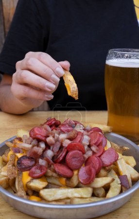 Photo for Closeup view of a woman holding a fried potato dipped in cheddar cheese sauce. The french fries with sausages and bacon dish besides a glass of beer. - Royalty Free Image