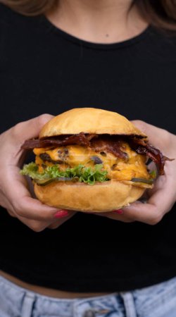 Foto de Hamburguesa. Vista de cerca de una mujer sosteniendo una hamburguesa con carne a la parrilla, lechuga, pepino encurtidos, queso cheddar y tocino crujiente. - Imagen libre de derechos